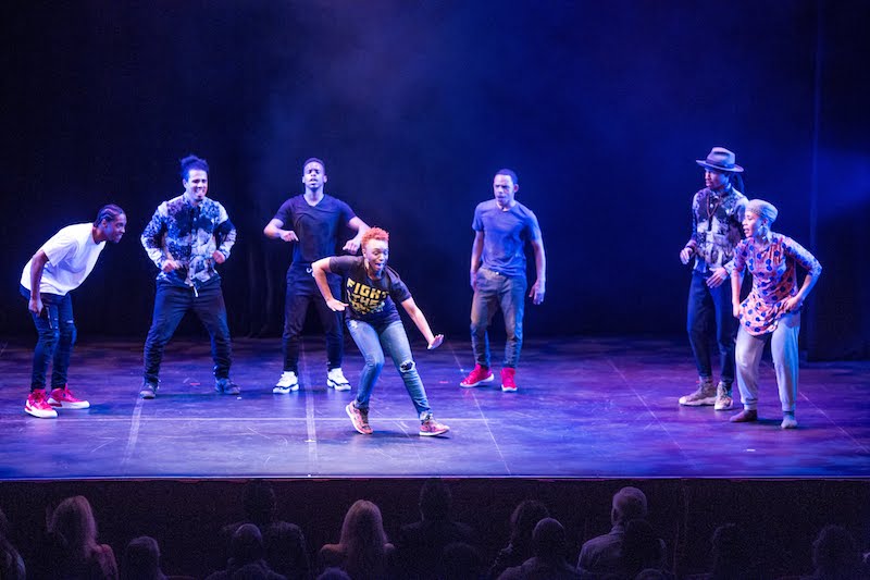 A woman improvs in jeans, a t-shirt and red sneakers. A group of performers cheer her on.